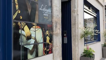La façade d’un hôtel ornée d’une grande enseigne publicitaire représentant des personnages historiques, d’une porte bleue et d’un petit jardonier avec un arbre.
