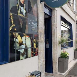 A hotel facade with a large advertisement featuring historical figures, a blue door, and a small planter with a tree. - Logis Hotel Lafayette (Rochefort)