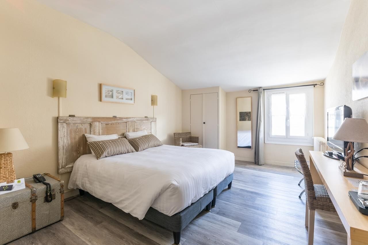 Un dormitorio amplio con una cama grande, un escritorio y una silla. Hay una maleta en el suelo y una lámpara sobre el escritorio.