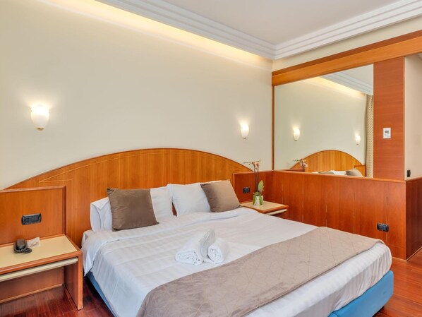 Habitación de hotel con cabecera de madera, un espejo grande, mesita de noche con teléfono y una cama bien hecha con toallas.