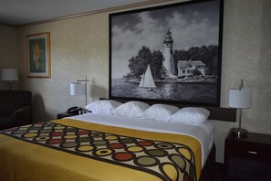 Ein Hotelzimmer mit einem Bett, zwei Nachttischlampen, einem Schreibtisch mit Telefon, einem Sessel und einem großen gerahmten Bild eines Leuchtturms an der Wand.