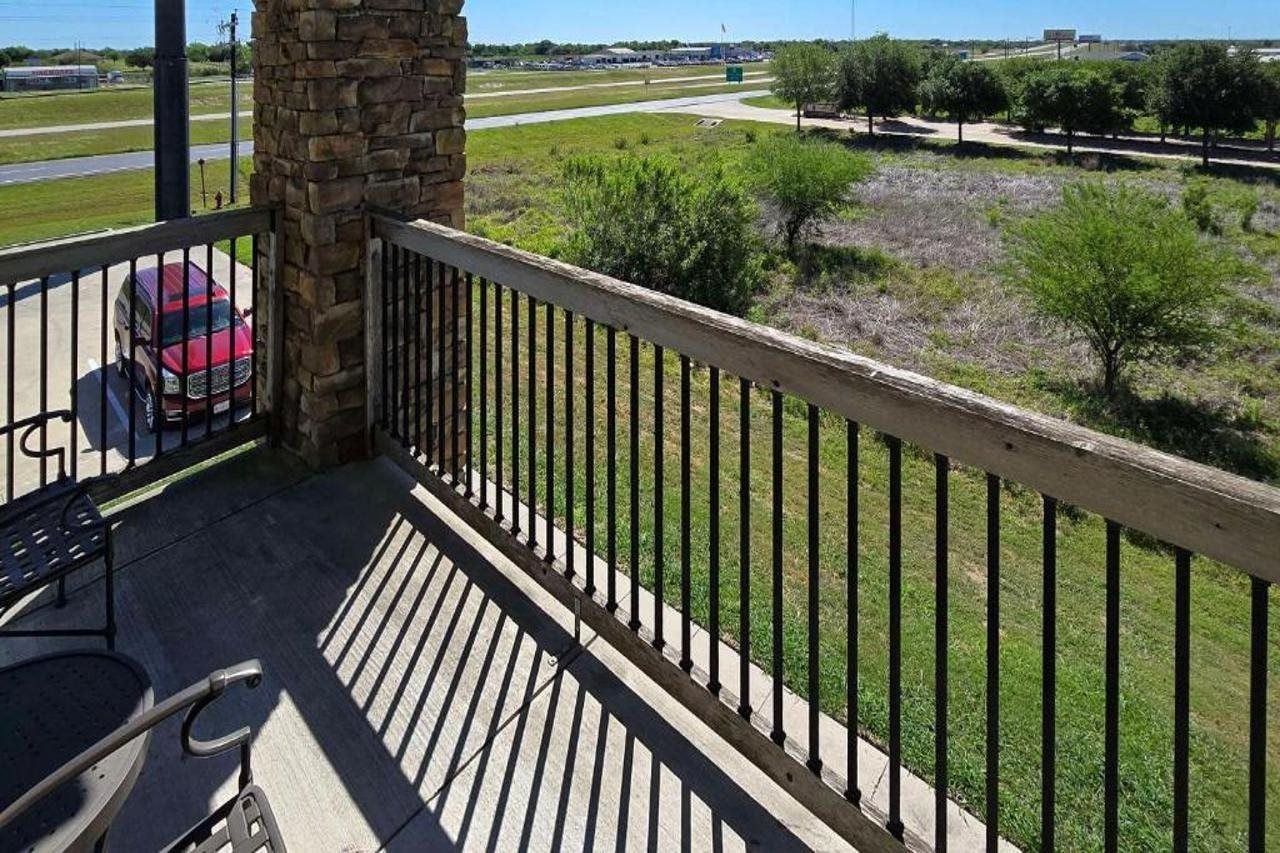 A balcony with a stone pillar, a red vehicle parked outside, and a view of a grassy area with trees.