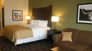 A hotel room with a bed, a nightstand, a lamp, a framed picture, a couch, and a wooden floor. - Yonder Inn (Beeville)