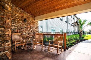 Un patio techado con mecedoras de madera y una mesa, pared de piedra, y vistas a un edificio con amplias ventanas.