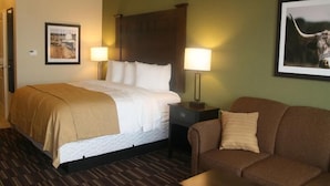 A hotel room with a bed, a nightstand, a lamp, a framed picture, a couch, and a wooden floor. - Yonder Inn (Beeville)