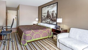 A hotel room with a bed, a desk, a chair, a sofa, and a large cityscape painting on the wall. - Delavan Inn and Suites (Delavan)