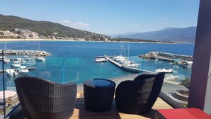 A balcony with wicker chairs, a red table, and a view of a marina with boats and a beach. - Le Golfe, Piscine & Spa Casanera (Serra-di-Ferro)
