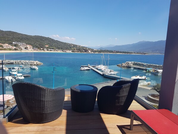 A balcony with wicker chairs, a red table, and a view of a marina with boats and a beach. - Hotel Le Golfe (Serra-di-Ferro)