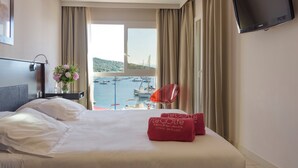 A hotel room with a bed, a TV, a vase with flowers, and a view of boats and a hill. - Le Golfe, Piscine & Spa Casanera (Serra-di-Ferro)