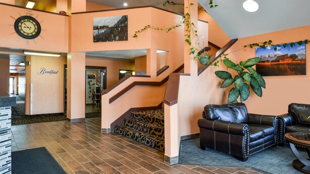 A lobby with a staircase, a clock, a couch, and a large plant. - Americinn by Wyndham Rawlins (Rawlins)