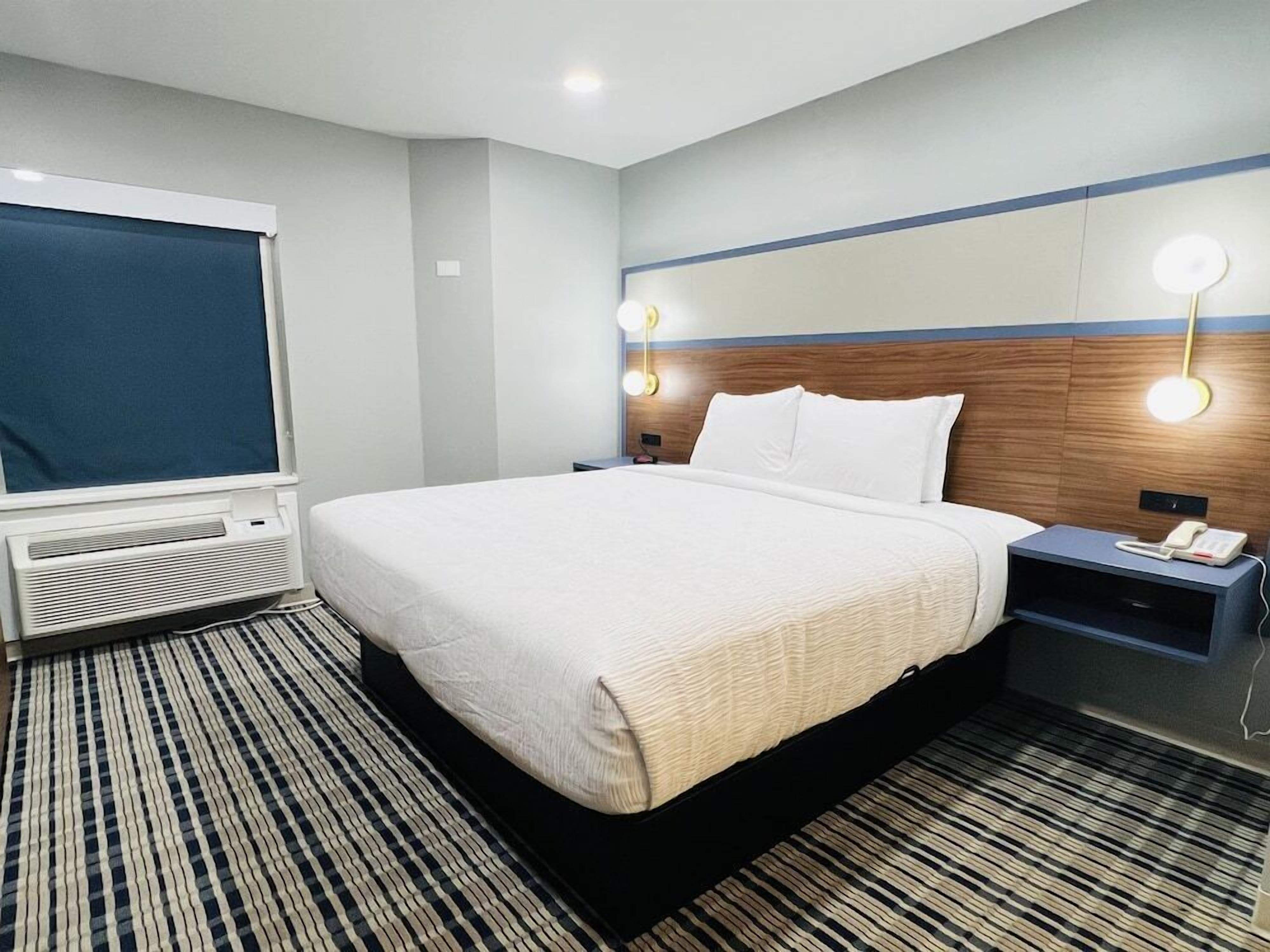 A hotel room with a bed, bedside tables, a lamp, a heater, and a window with a blind.