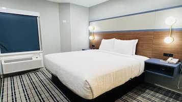 A hotel room with a bed, bedside tables, a lamp, a heater, and a window with a blind.