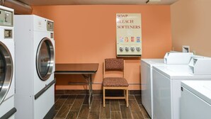 A laundry room with a washing machine, dryer, and a chair. There is a sign on the wall listing soap, bleach, and softeners. - Americinn by Wyndham Rawlins (Rawlins)