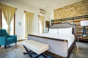 A hotel room with a large bed, a bench, a teal armchair, a bedside table with a lamp, and a brick-patterned wall. - Turtle Island Beach Resort Trademark Collection by Wyndham (San Pedro)