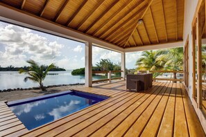Um deck de madeira com piscina, área coberta e vista para o mar e palmeiras.