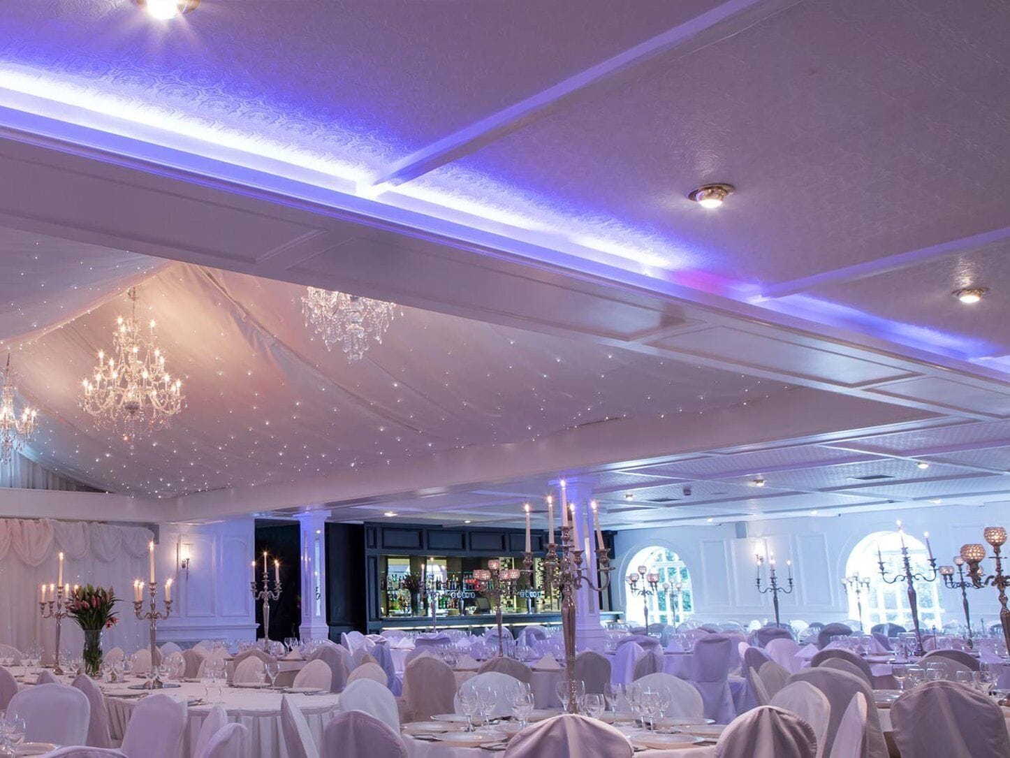 A banquet hall with round tables, chandeliers, and a bar area.