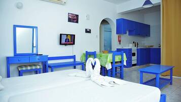 A hotel room with a bed, a desk, a chair, a TV, and a kitchenette in the background.