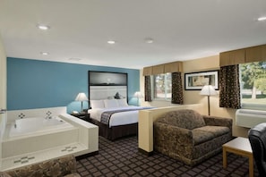 A hotel room with a large bathtub, a bed with a headboard, a sofa, a small table, and a window with curtains. - Super 8 by Wyndham Whitewater WI (Whitewater)