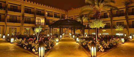 A hotel courtyard at night with a central gazebo, palm trees, and flower beds.