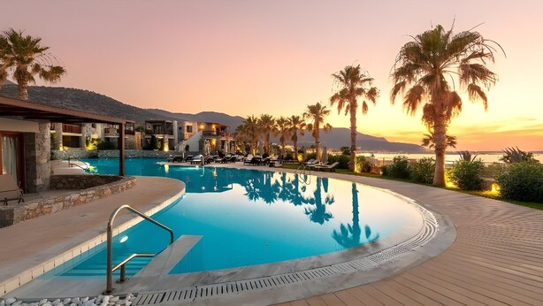 A pool area with lounge chairs, palm trees, and a view of the sea at sunset. - Ikaros Beach Luxury Resort and Spa - Adults Only (Hersonissos)
