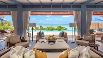 Amplia habitación con un gran ventanal con vistas a la playa, ventilador de techo y una mesa de centro con fruta fresca.