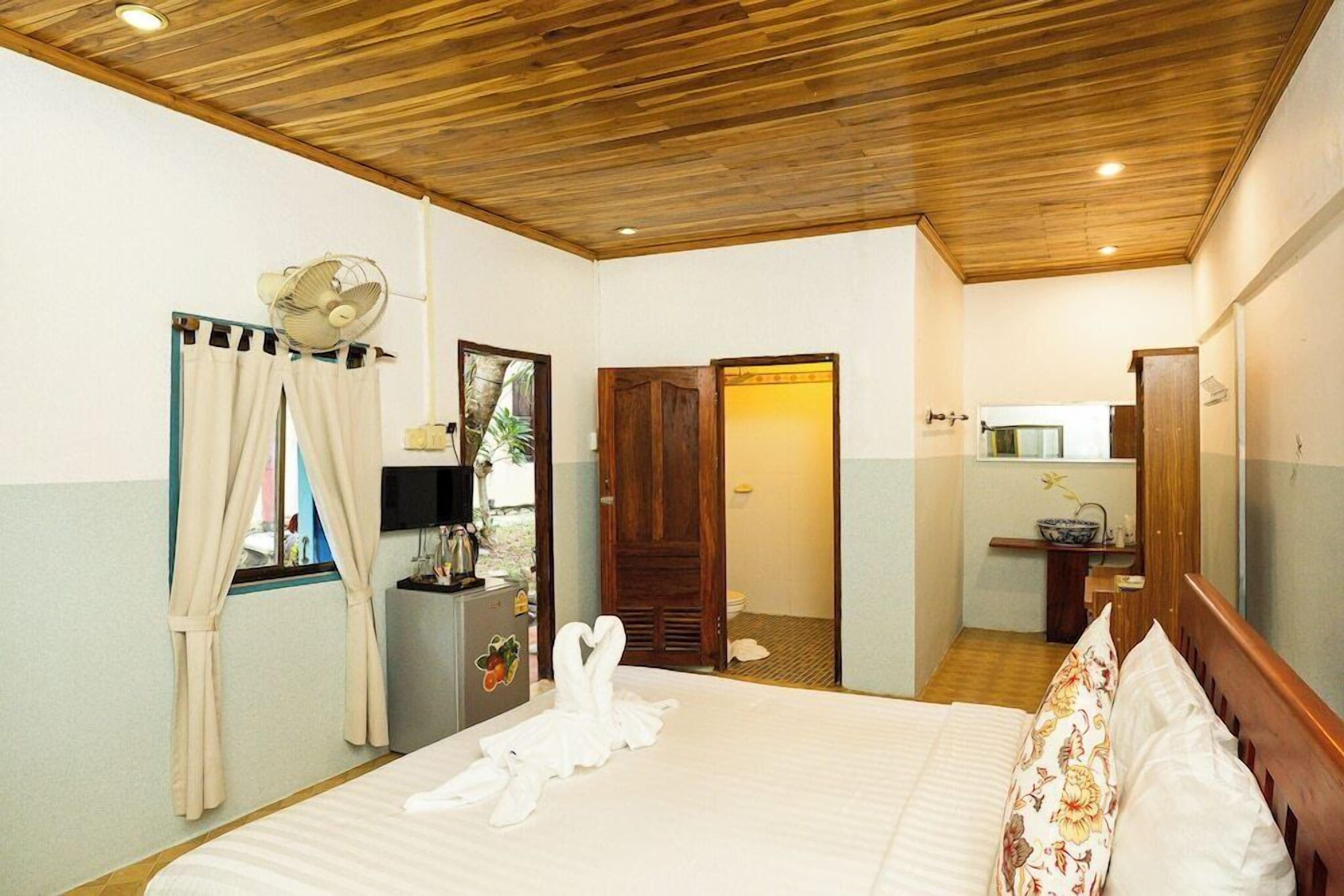 Una habitación de hotel con techo de madera, una cama con ropa blanca, un ventilador, un espejo, una ventana con cortinas y un baño pequeño.