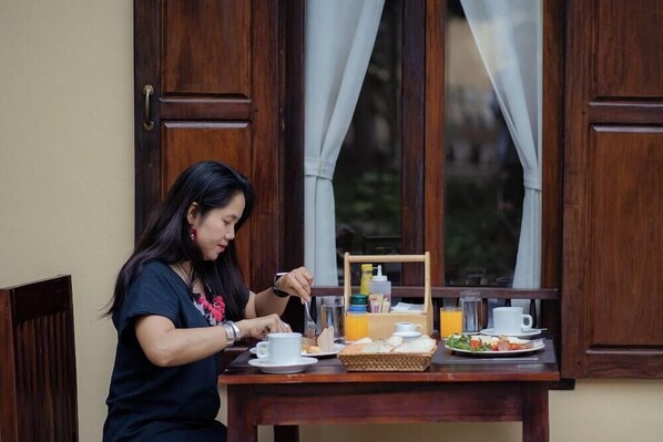A person dining at a wooden table with a window view, a basket of bread, a cup, and a glass of orange juice. - Khoum Xieng Thong Boutique Villa (Luang Prabang)