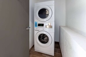 A front-loading washing machine with a digital display and control panel, located in a hallway with a door and a bench. - Microtel Inn & Suites by Wyndham Sunbury/columbus North (Sunbury)