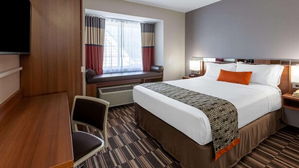 A hotel room with a bed, a desk, a chair, a TV, a window with curtains, and a nightstand with a lamp.