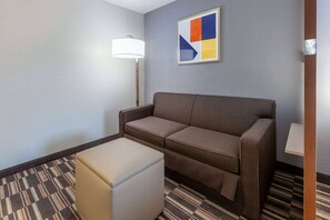 A hotel room with a sofa, a small ottoman, a floor lamp, and a wall art piece. - Microtel Inn & Suites by Wyndham Sunbury/columbus North (Sunbury)