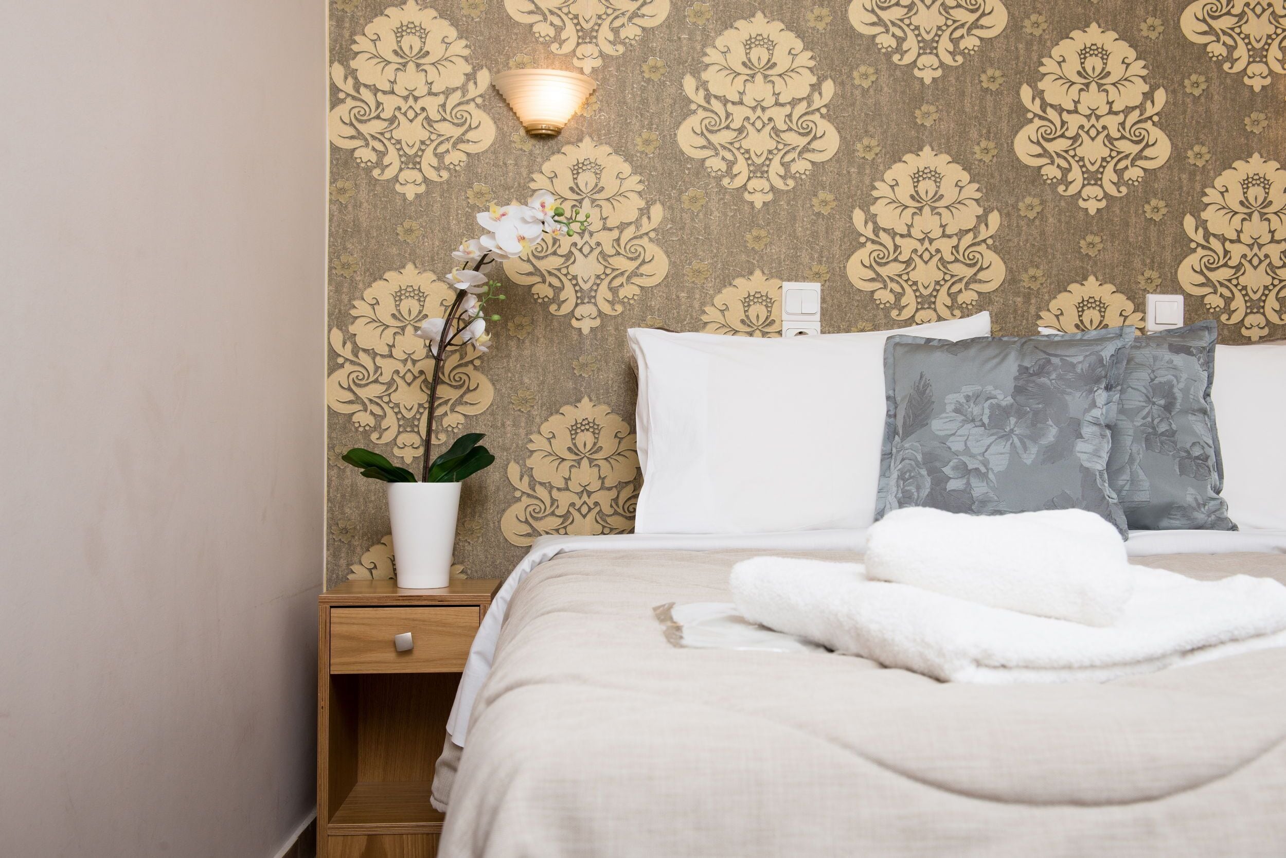 Una cama bien hecha con almohadas blancas y grises, un jarrón blanco con una planta, y una pared con papel tapiz estampado.