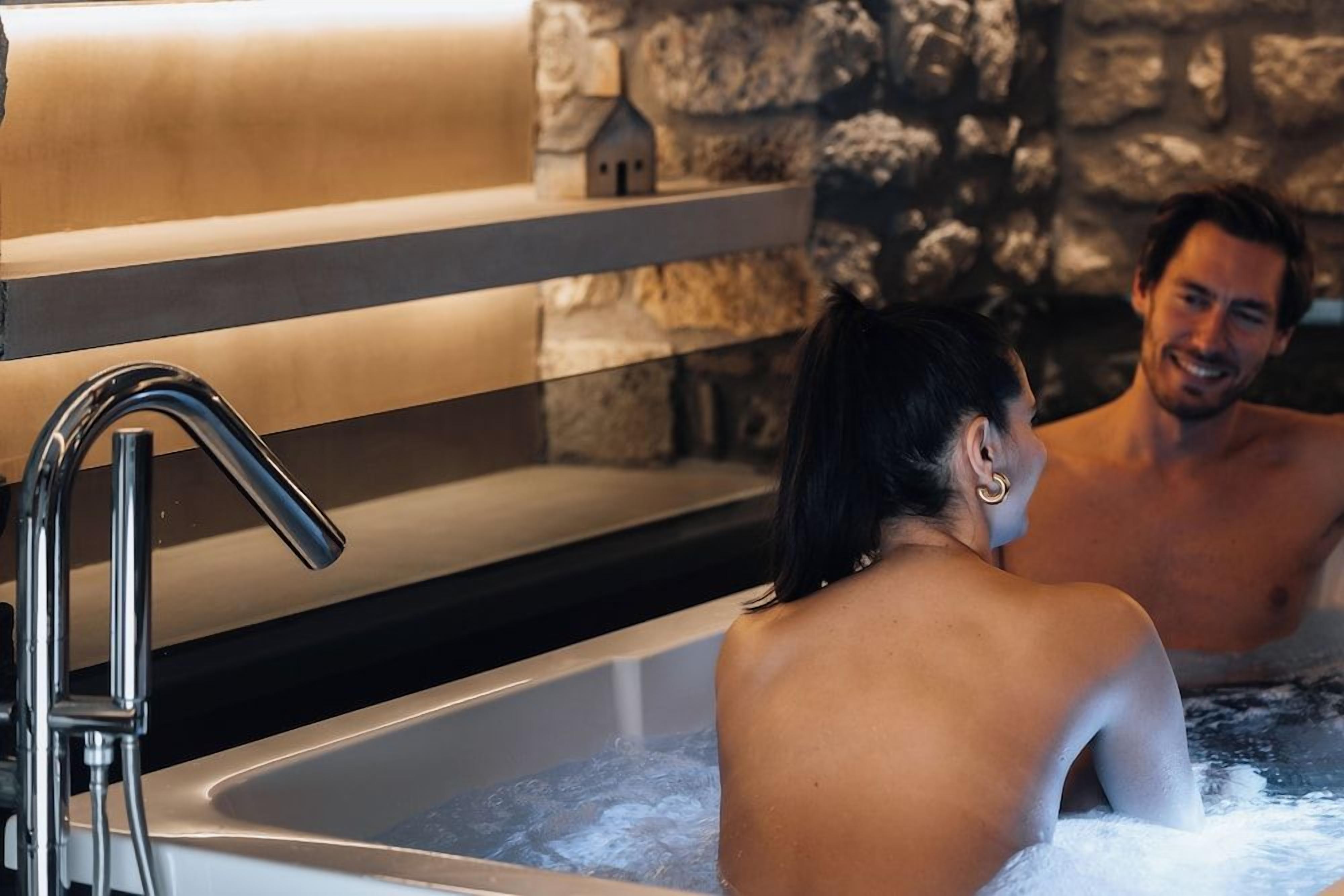 A bathtub with a stone wall backdrop and a small house model on a shelf.