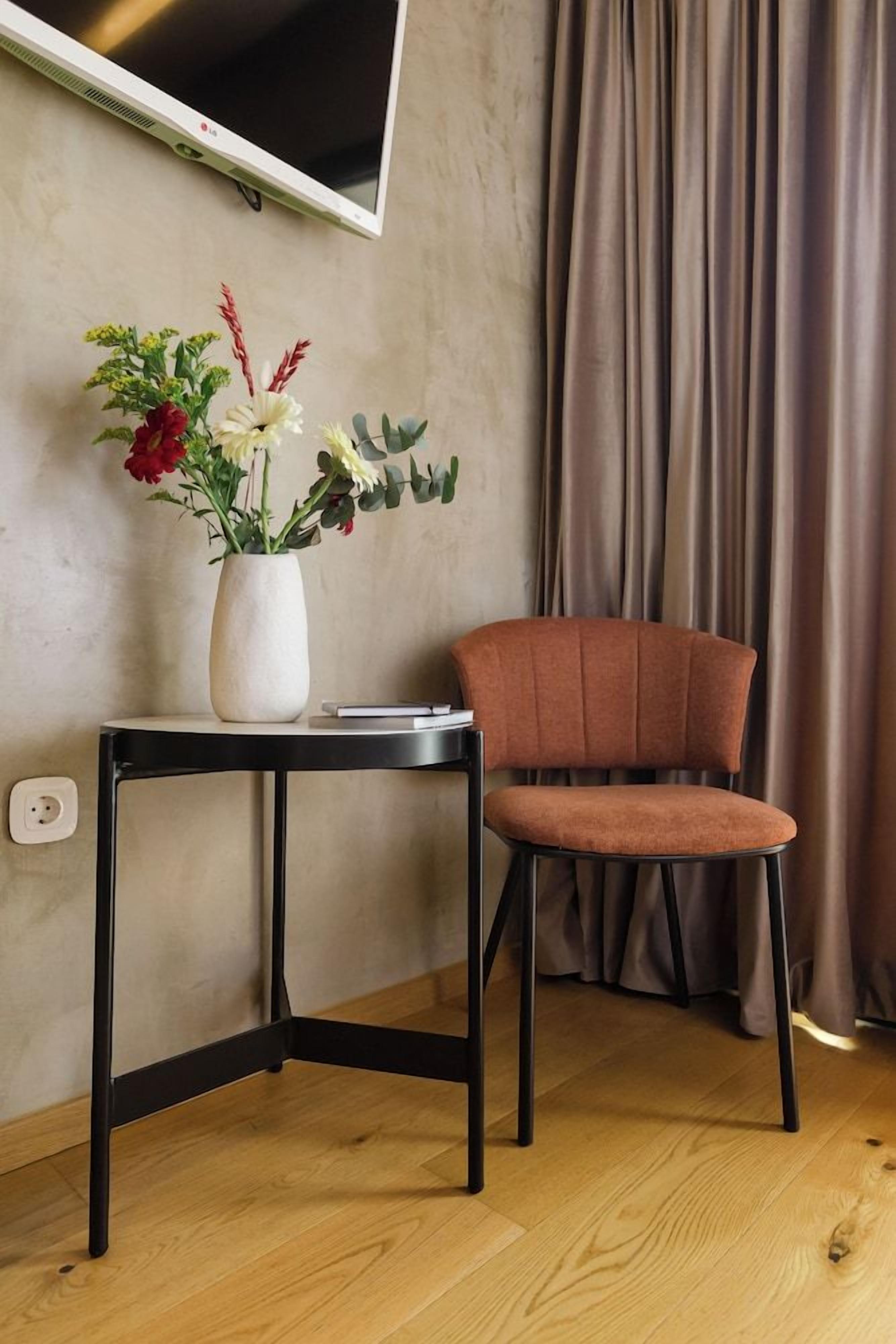 A room with a television mounted on the wall, a small table with a vase of flowers, a brown chair, and a curtain.