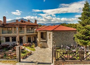 Un edificio de piedra con techo de tejas rojas, asientos al aire libre y montañas al fondo.