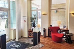 A hotel lobby with a reception desk, a red chair, a small table with a lamp, and a large window. - Select Hotel Tiefenthal (Hamburg)