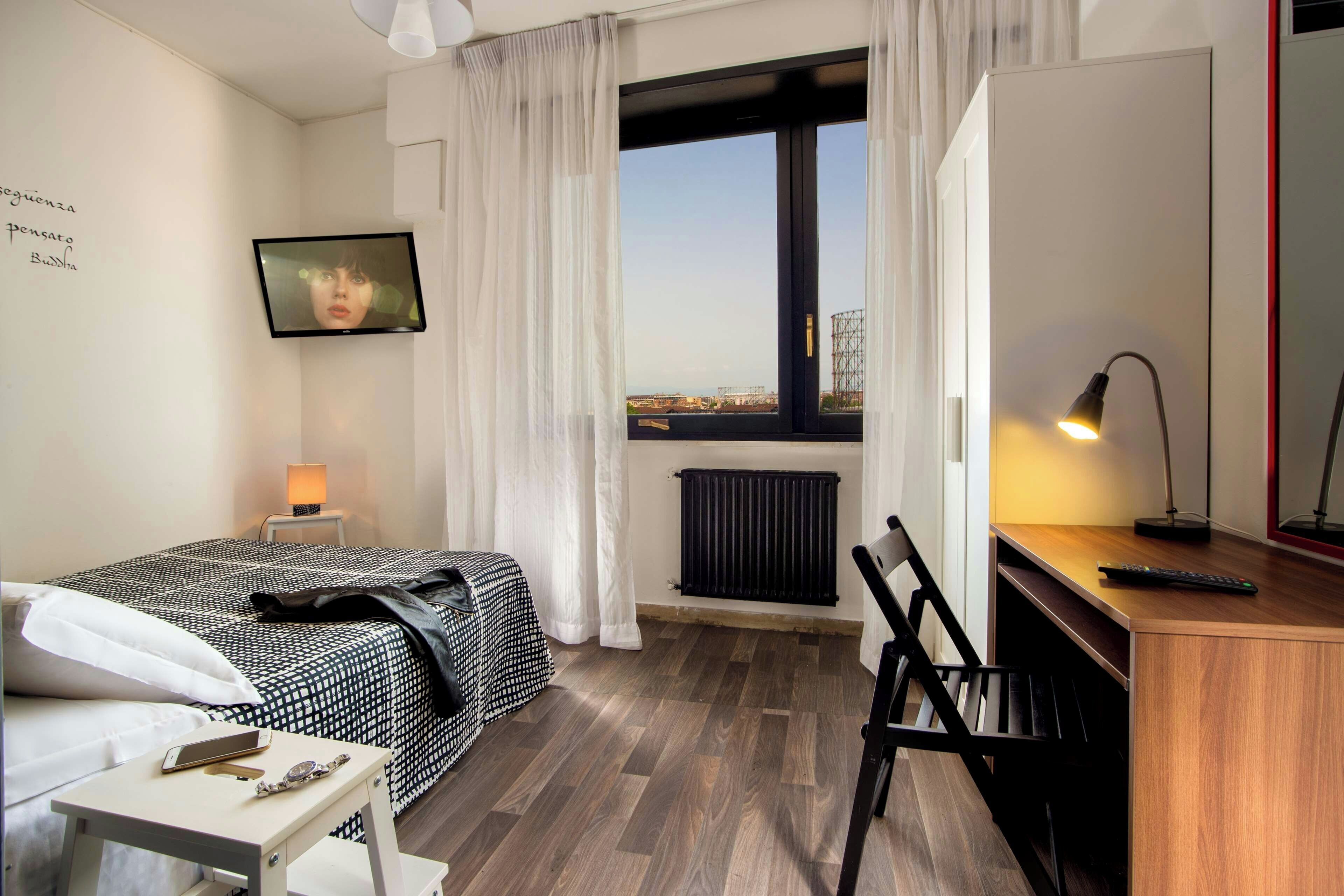 A hotel room with a bed, a desk, a chair, a TV, and a view of the cityscape through the window.