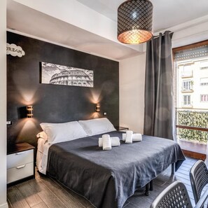 A modern hotel room with a bed, two bedside lamps, a large window with a view, and a wall-mounted picture of the Colosseum. - Radio Hotel (Rome)
