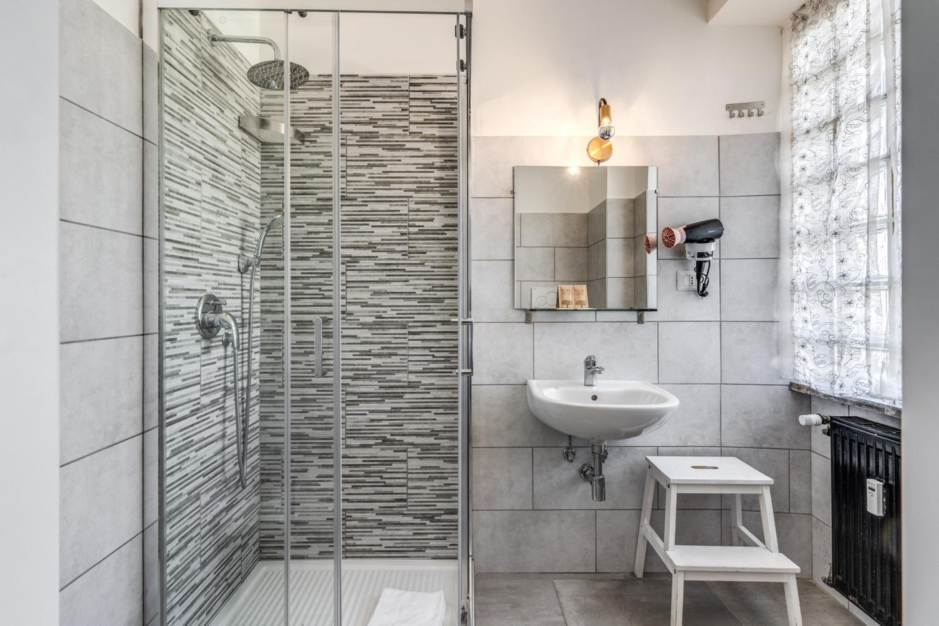 Un baño moderno con cabina de ducha de vidrio, un lavabo blanco, un taburete y un secador de pelo montado en la pared.