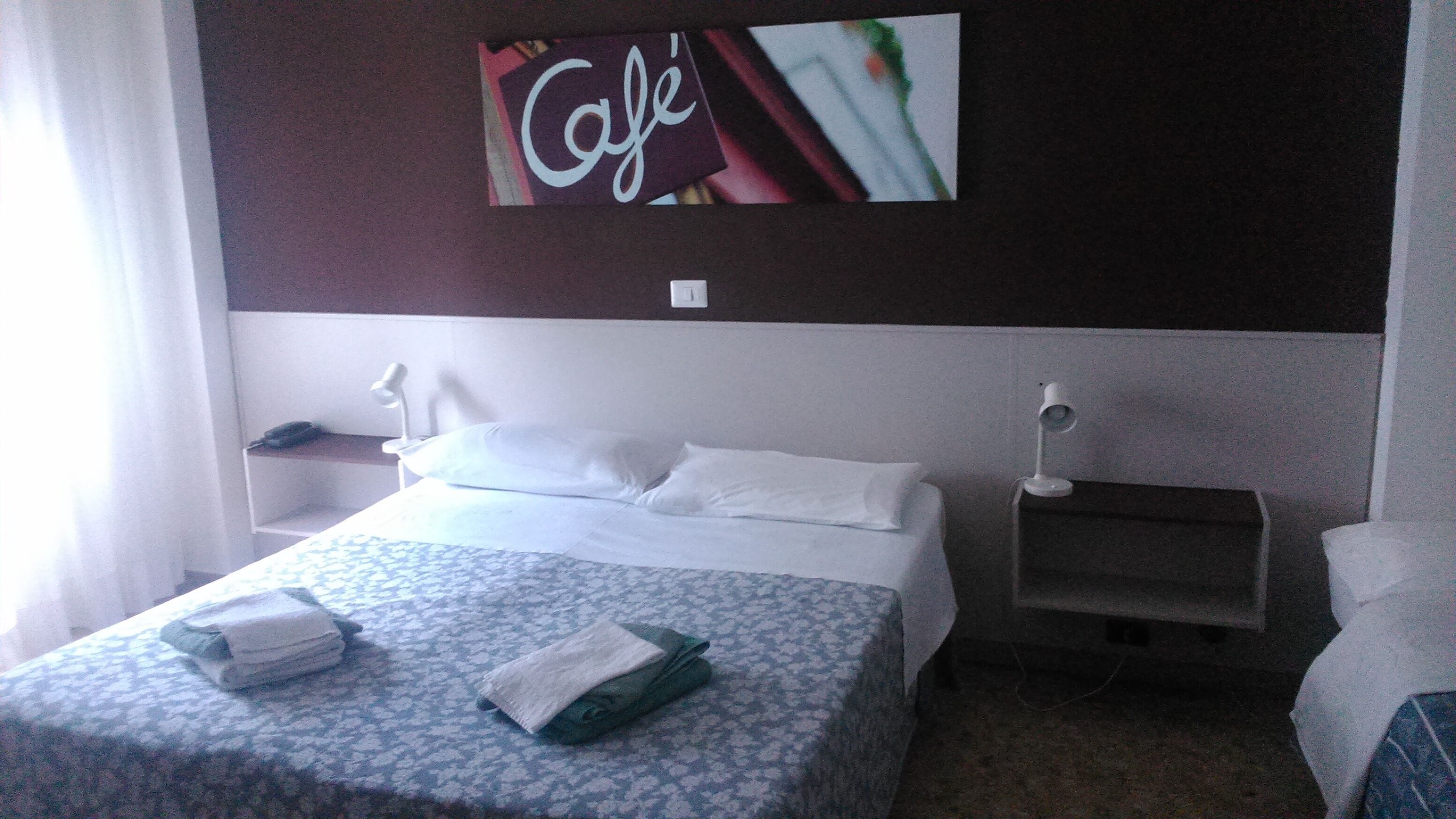 A hotel room with a bed, a nightstand, a lamp, and a wall with a 'Café' sign.