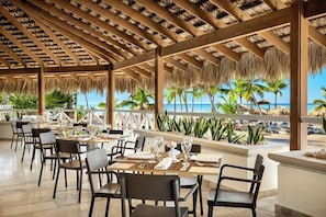 A covered outdoor dining area with tables and chairs, a view of the beach, and palm trees. - Sunscape Dominicus La Romana (San Rafael del Yuma)