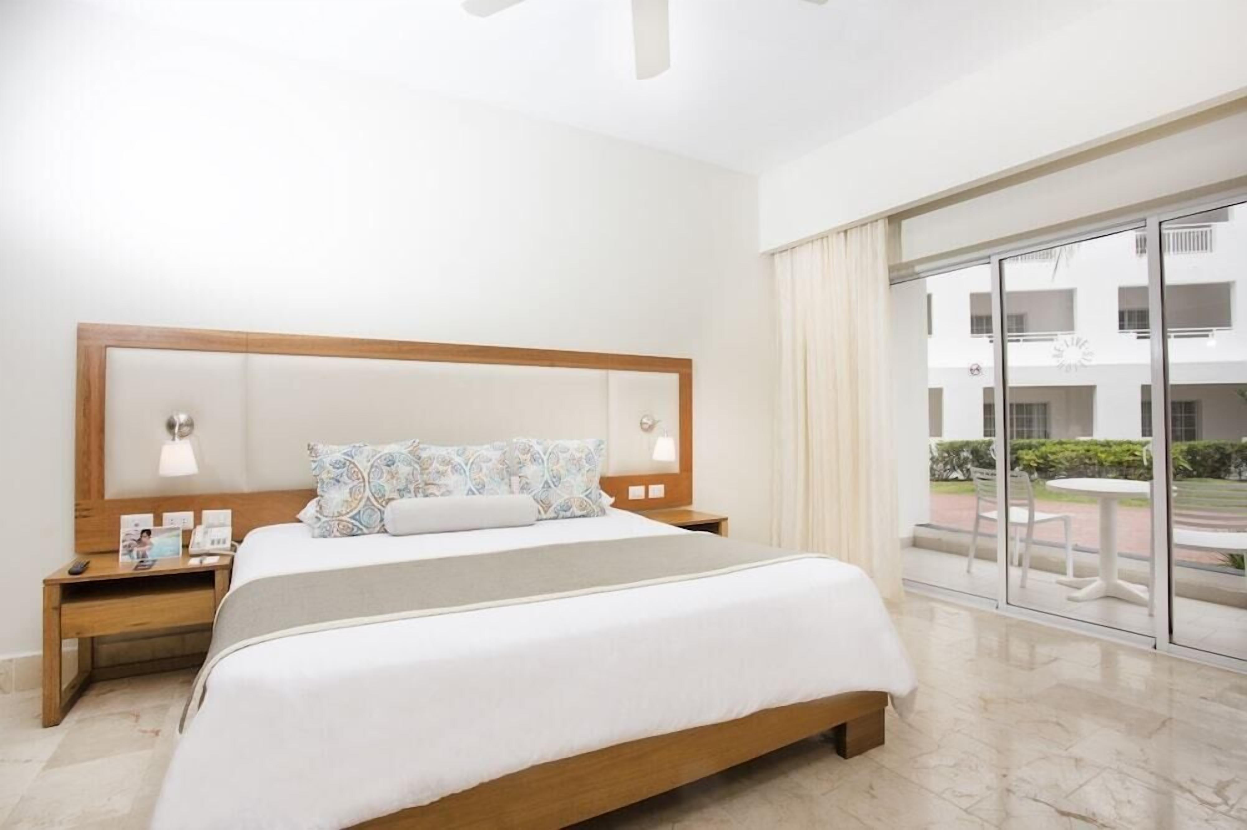 A hotel room with a large bed, a nightstand with a lamp, a window with a view of the outside, and a balcony with a table and chairs.