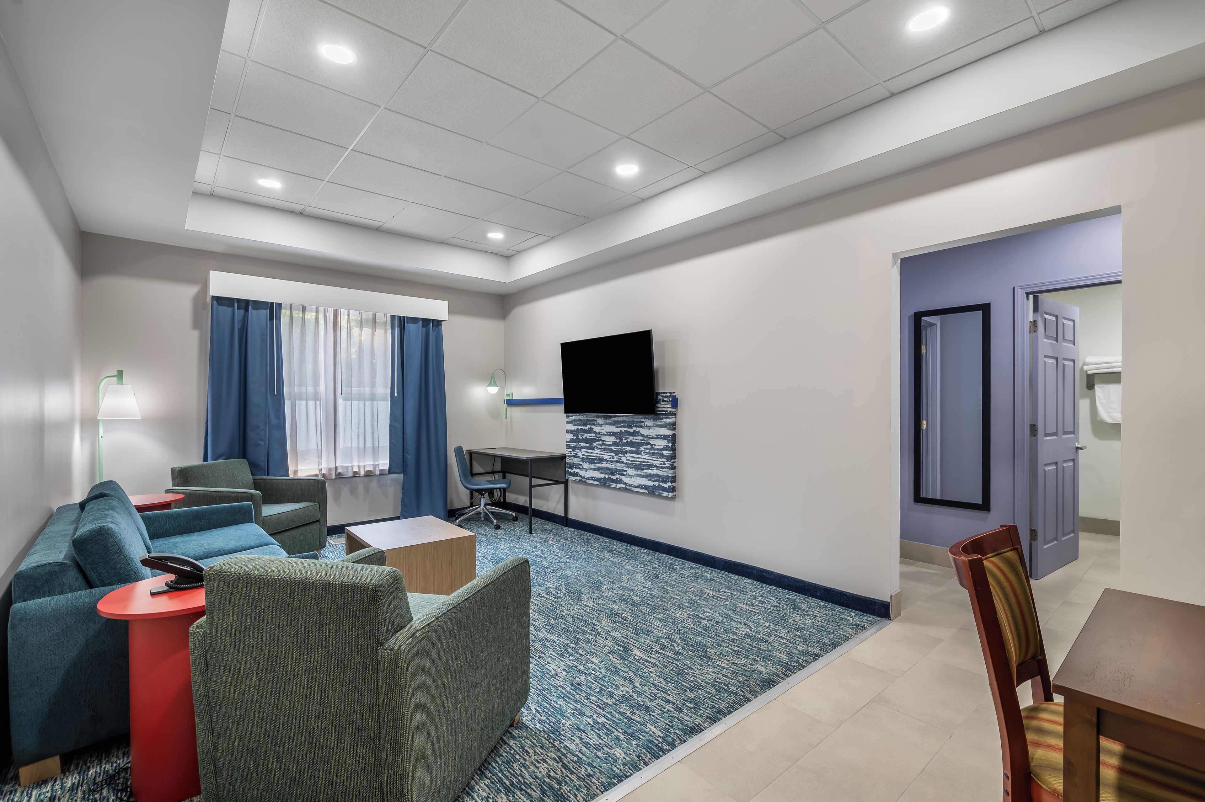 A modern hotel room with a sofa, armchair, desk, and a flat-screen TV mounted on the wall.