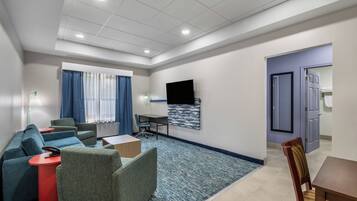 A modern hotel room with a sofa, armchair, desk, and a flat-screen TV mounted on the wall.
