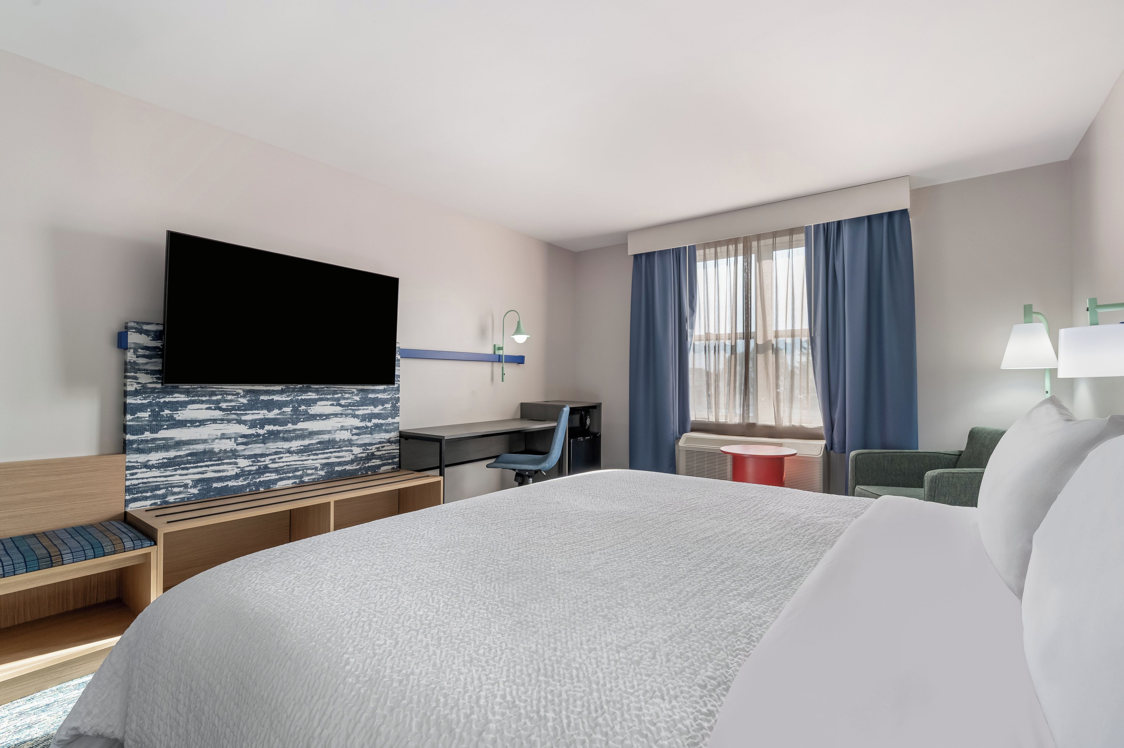 A hotel room with a large bed, a desk, a chair, a TV, and a window with curtains.