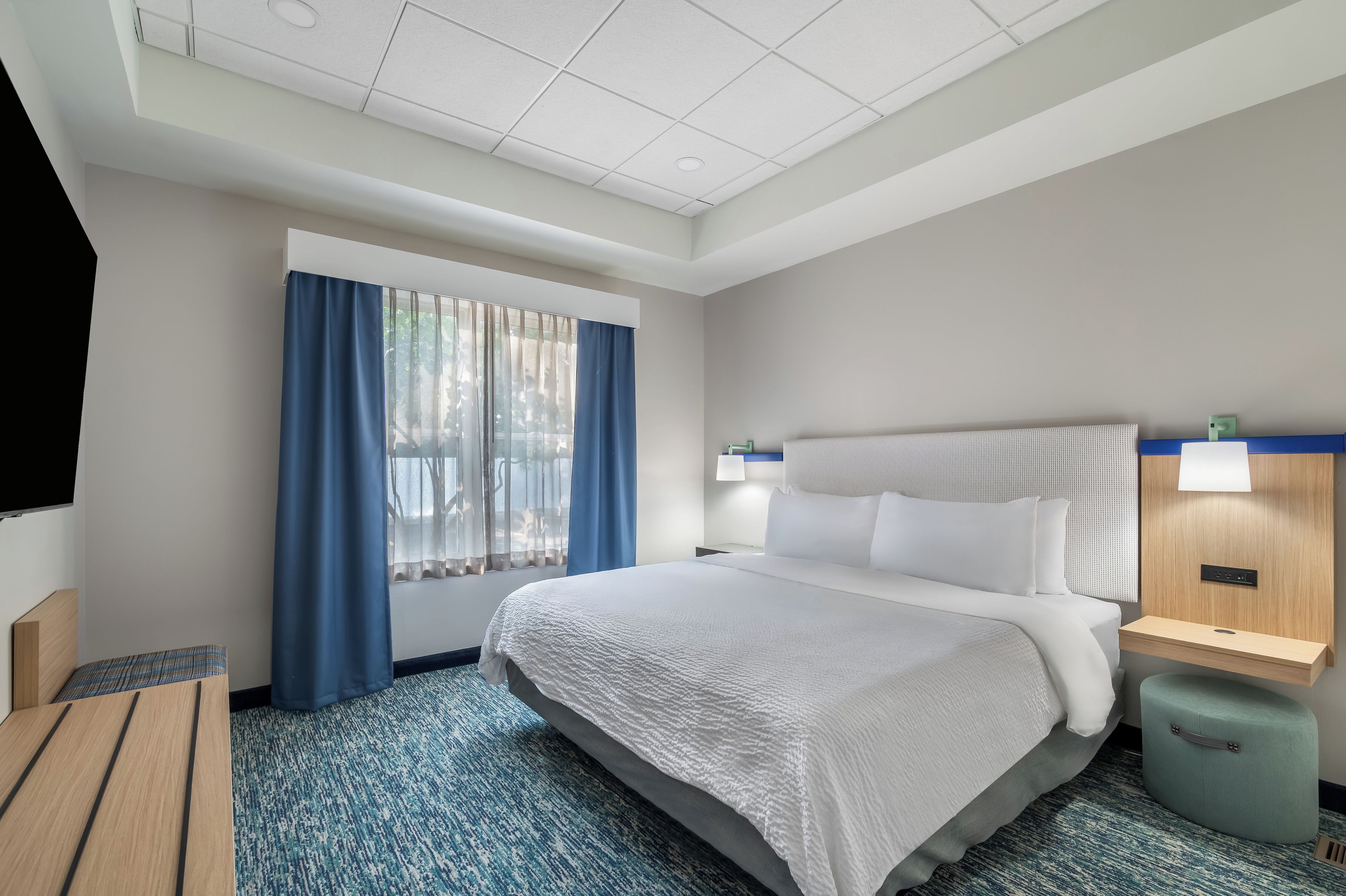 A hotel room with a large bed, a desk, a chair, a TV, and a window with curtains.