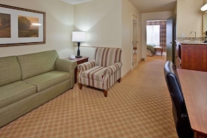 A hotel room with a green sofa, a striped armchair, a small table with a lamp, a wooden desk, and a bathroom visible through an open door. - Country Inn & Suites by Radisson, Birch Run-frankenmuth, MI (Birch Run)