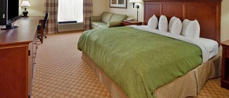 A hotel room with a large bed, a desk with a computer, a chair, a bedside table with a lamp, and a window with curtains.