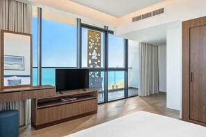 A modern hotel room with a large TV, wooden furniture, a balcony with a view of the sea, and large windows.