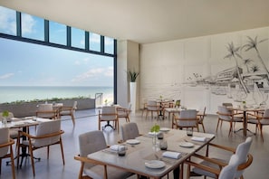 A dining area with tables and chairs, large windows, and a view of the sea.