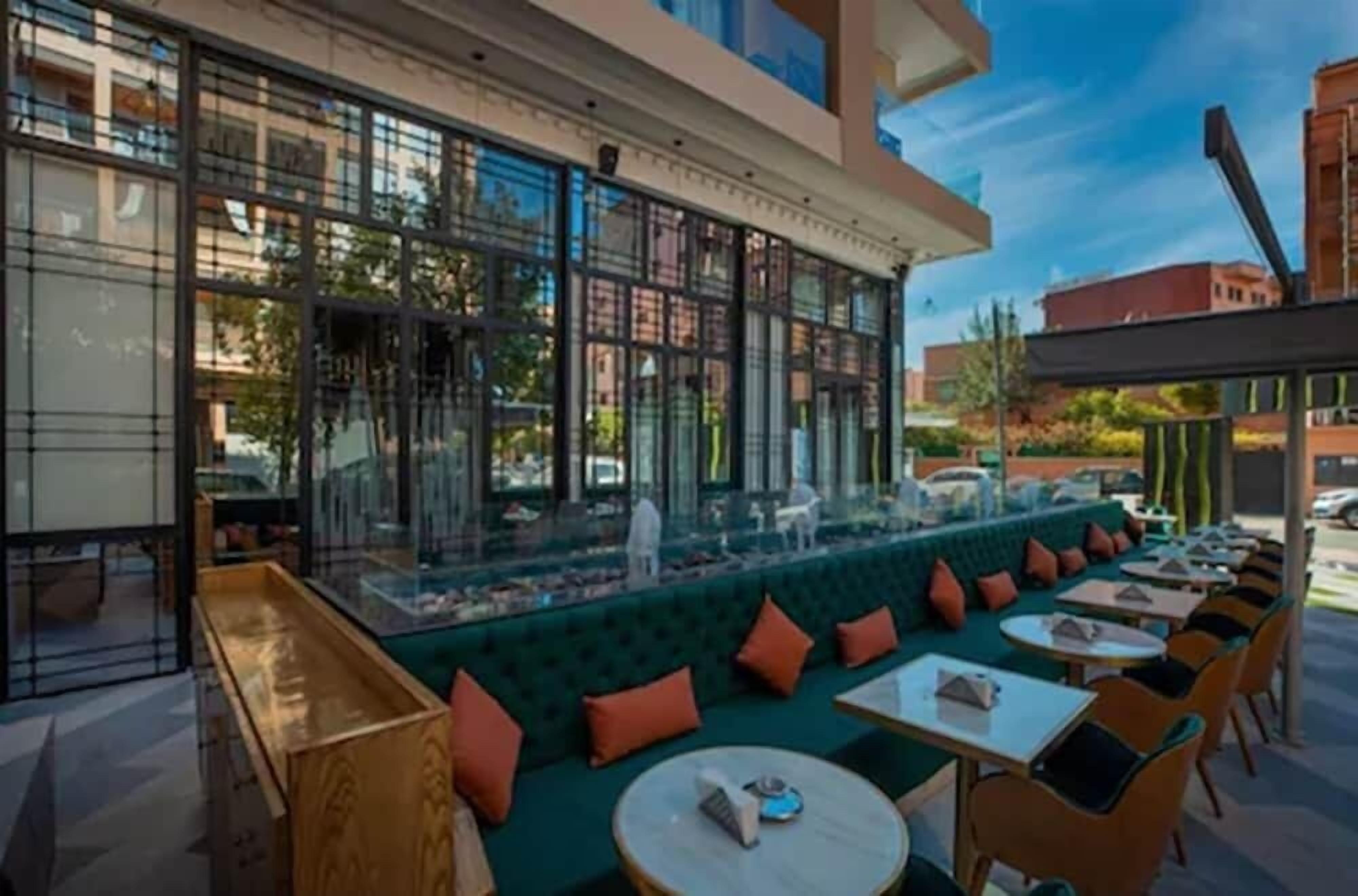 Un espace extérieur aménagé avec des tables et des chaises, un banc vert avec coussin, et une structure en verre offrant une vue sur la rue.